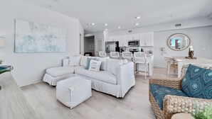 A modern living room with a sectional sofa, a coffee table, and a wicker chair. There is a kitchen area with white cabinets and a dining table.