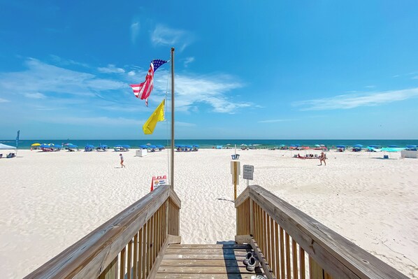 A wooden boardwalk leading to a beach with flags, umbrellas, and beachgoers.