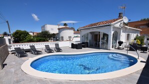 A swimming pool with lounge chairs, a covered seating area, and a white building with a tiled roof.