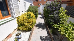 A narrow pathway flanked by potted plants and a building with a white exterior and grey shutters.