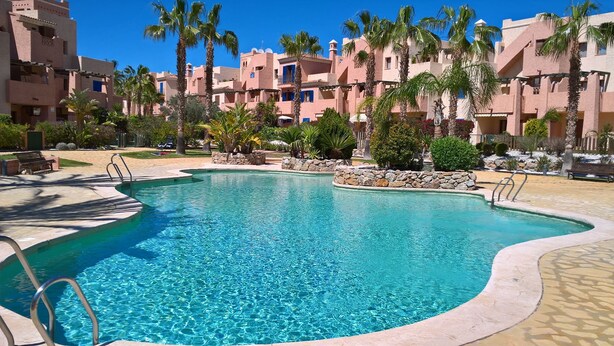 A swimming pool with clear blue water, surrounded by palm trees and a stone wall. - Attic apartment in Vera Beach with beautiful sea and mountain views (Vera Playa)