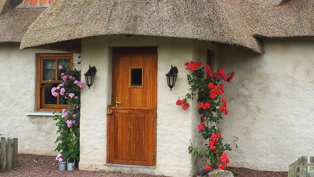 A thatched-roof cottage with a wooden door, two windows, and a small porch area with potted plants.