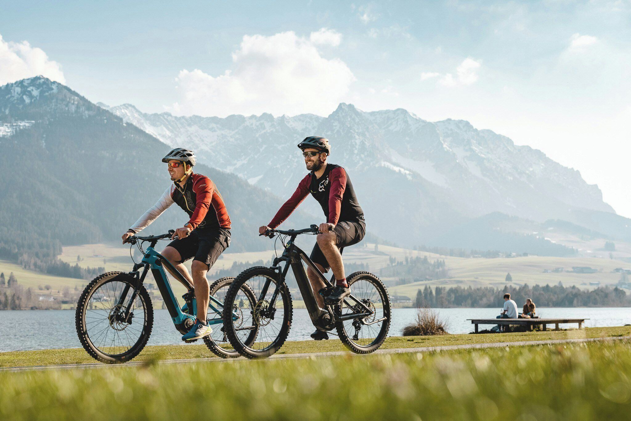 Two cyclists on a path with a lake and mountains in the background.