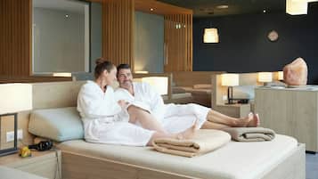 A couple in white robes on a bed in a modern room with wooden paneling, a clock on the wall, and a large rock on a side table.