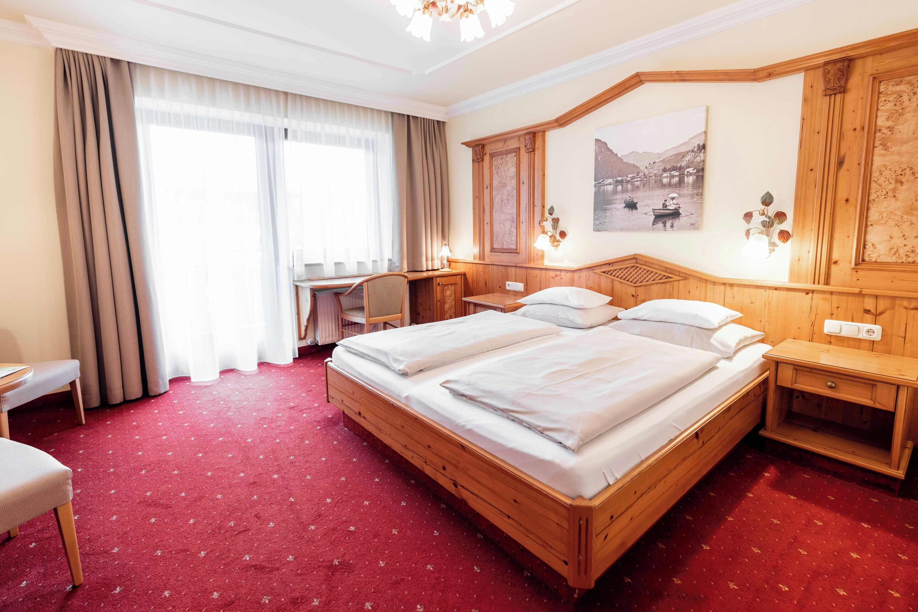 Ein Hotelzimmer mit einem großen Bett, einem Schreibtisch, einem Sessel und Blick auf einen See durch das Fenster.