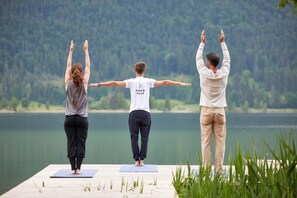 Three people practicing yoga on a wooden dock by a lake with a forested background. - Das Walchsee Resort (Walchsee)