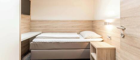 Habitació compacte i moderna amb un llit individual, una taula de nit i un reposabits integrat.