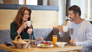Dos personas sentadas en una mesa con alimentos para el desayuno, incluyendo fruta y un vaso de jugo de naranja.