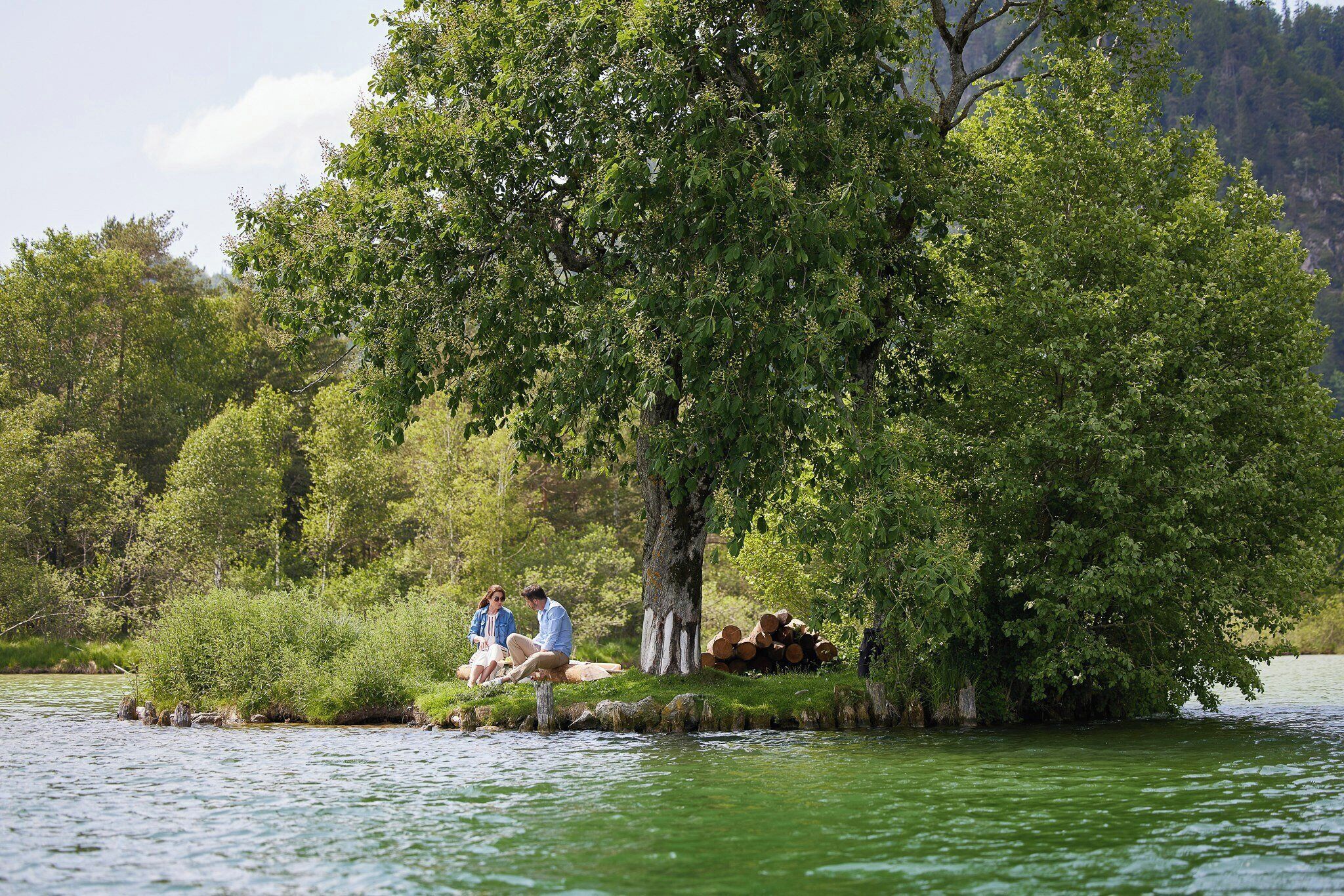 Una pareja sentada junto a un lago, rodeada de abundante vegetación y árboles.