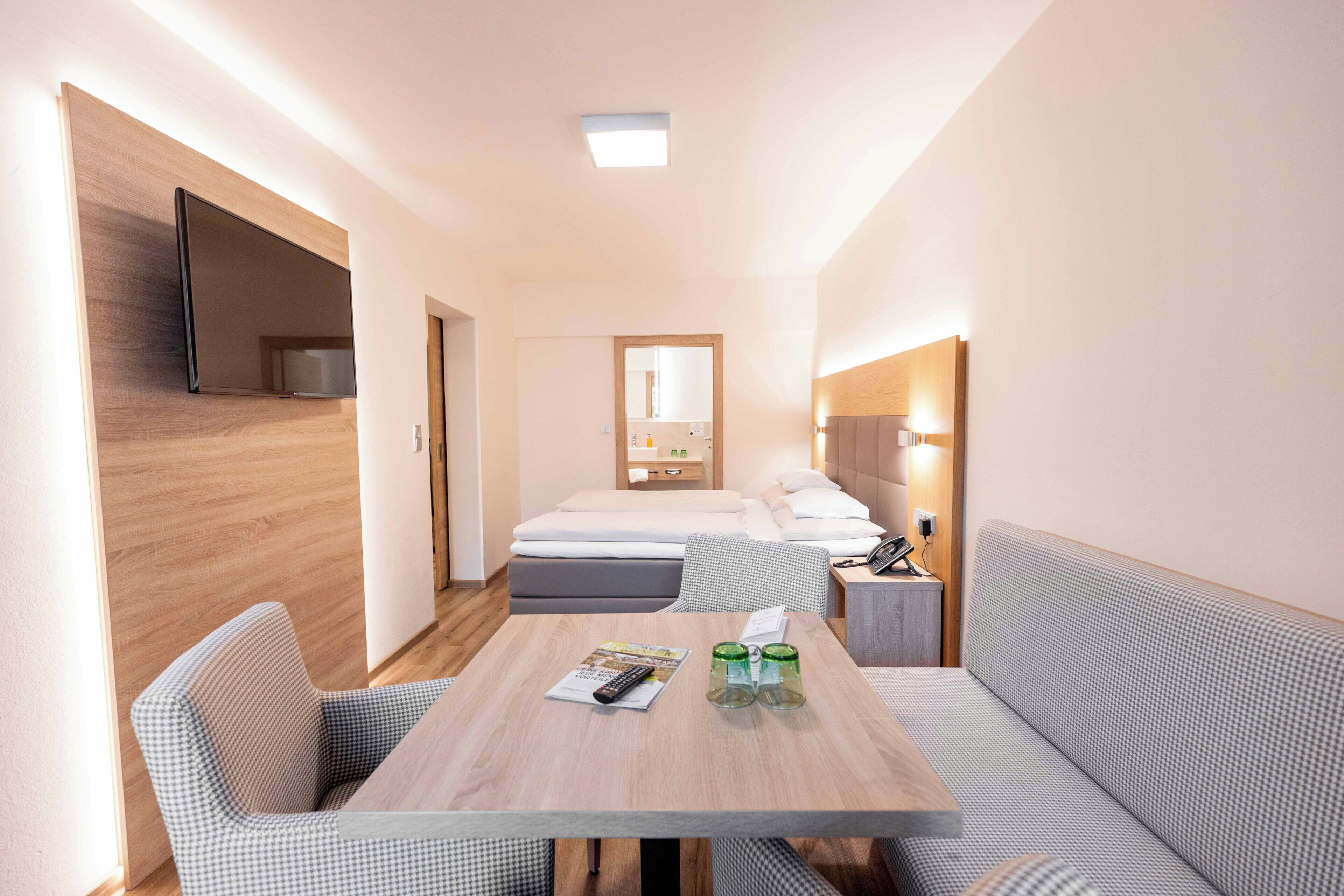 A hotel room with a bed, a bedside table with a lamp, a TV mounted on the wall, and a seating area with a table and chairs.