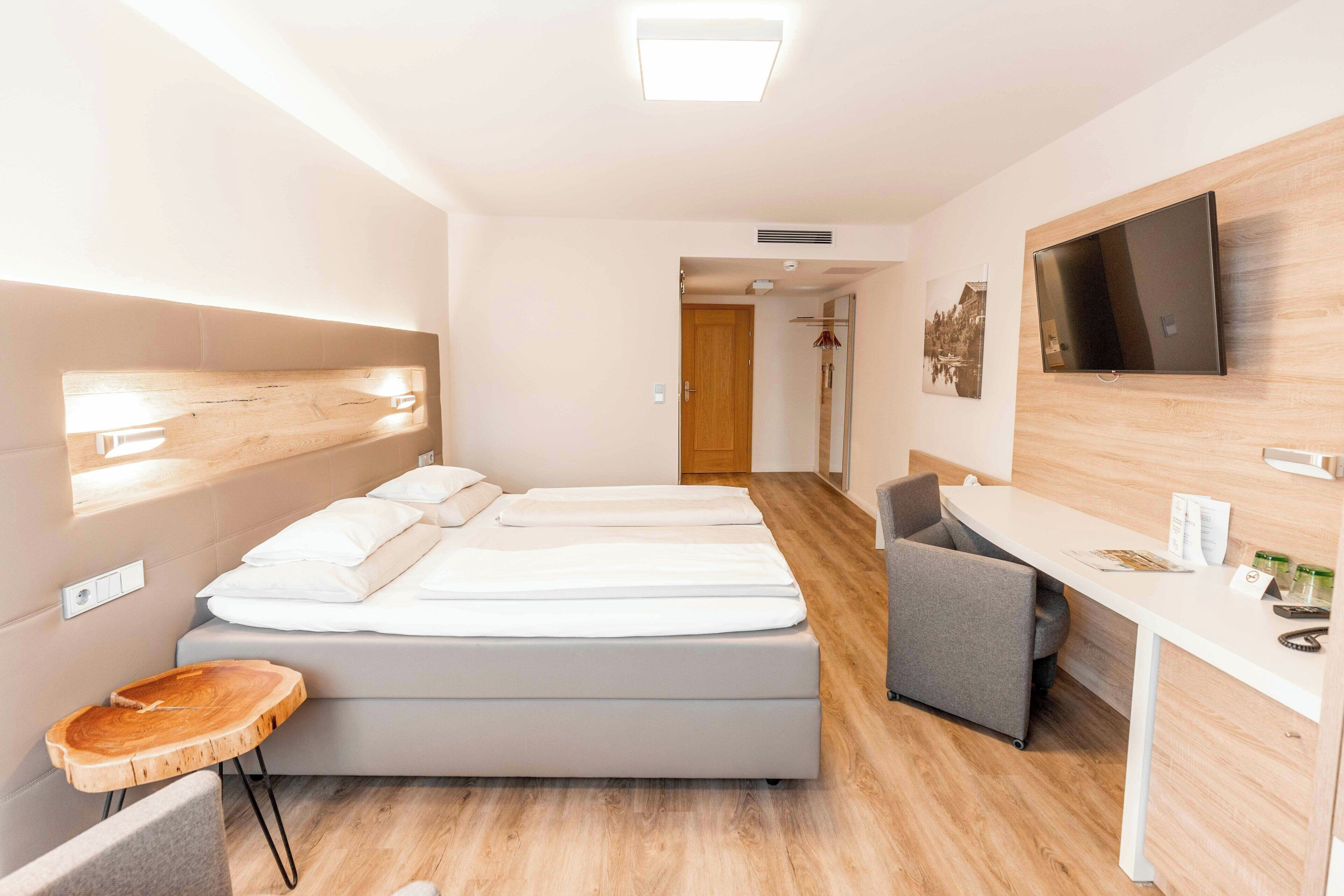 A hotel room with a bed, a desk, a chair, a TV, and a wooden headboard.