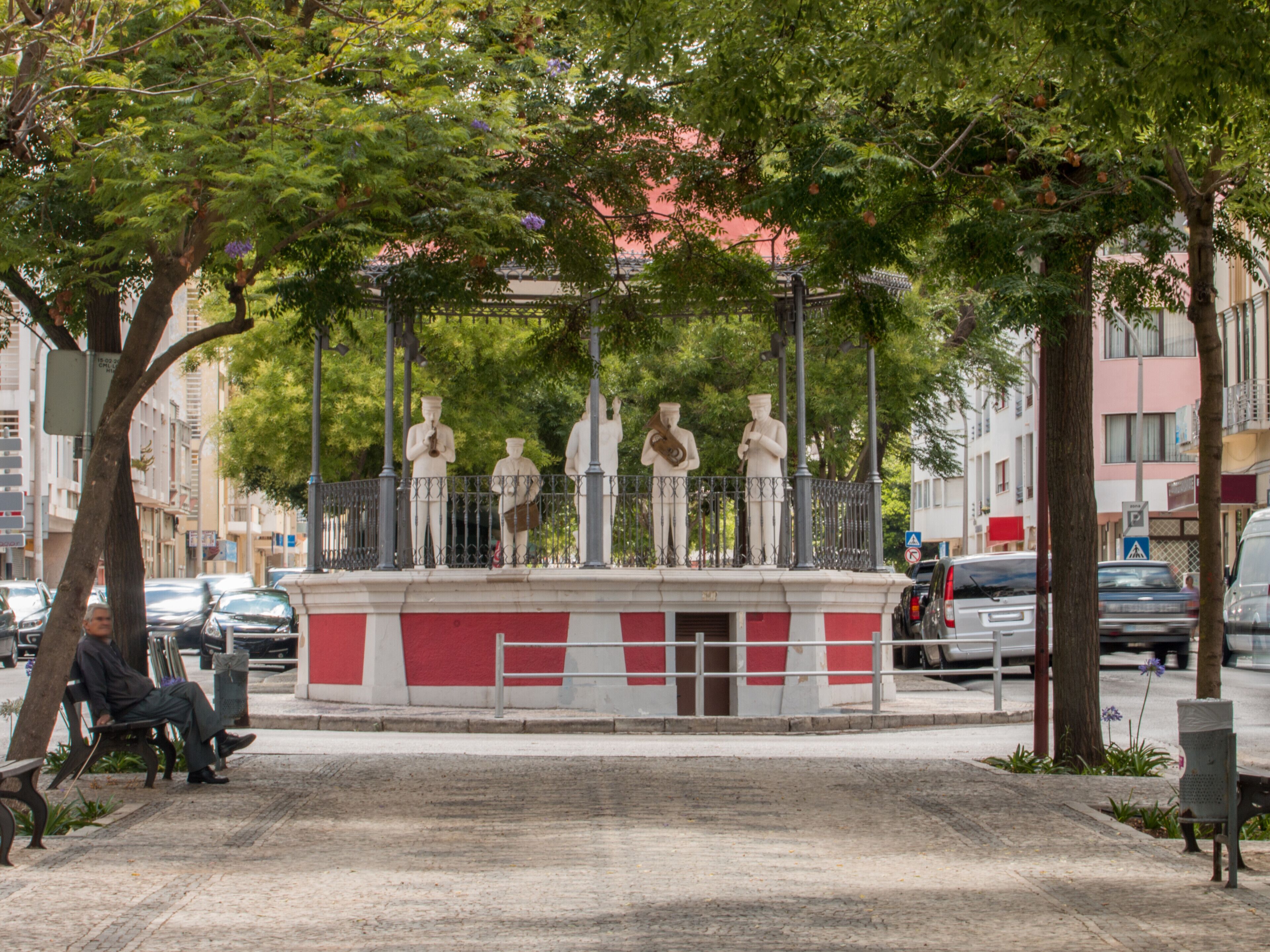 A public square with a central statue, benches, and surrounding trees.