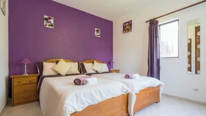 A double bed with white linens, a wooden headboard, and a purple wall. There are two bedside tables with lamps, a window with purple curtains, and a mirror on the wall.