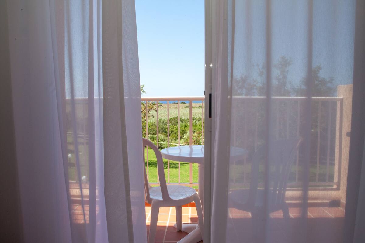 Ein Balkon mit weißem Tisch und Stühlen, Blick auf eine grüne Rasenfläche und einen strahlend blauen Himmel.