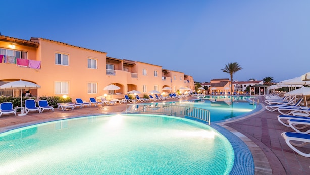 A swimming pool with lounge chairs and umbrellas, surrounded by buildings with balconies and windows. - Arenas de Son Bou (Alayor)