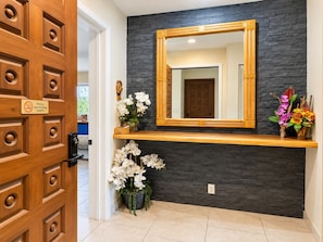 A wooden door with a non-smoking sign, a mirror above a dark stone wall, and a shelf with white and purple flowers.