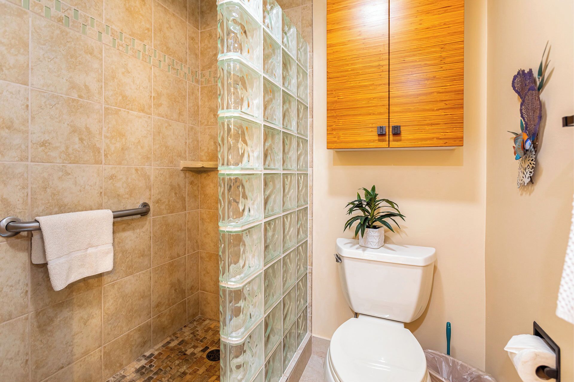 A bathroom with a glass shower enclosure, a wooden cabinet above the toilet, and a potted plant on the floor.