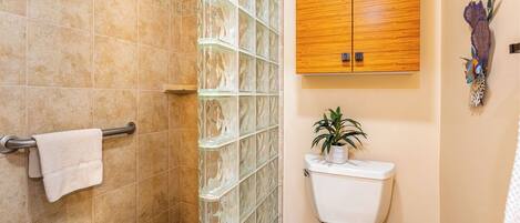 A bathroom with a glass shower enclosure, a wooden cabinet above the toilet, and a potted plant on the floor.