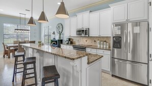 Una cocina moderna con gabinetes blancos, encimera de granito, electrodomésticos de acero inoxidable e iluminación colgante.