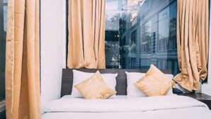 A bed with white bedding and two beige pillows, a window with beige curtains, and a view of buildings outside. - Hotel Suraj Palace (Bhopal)