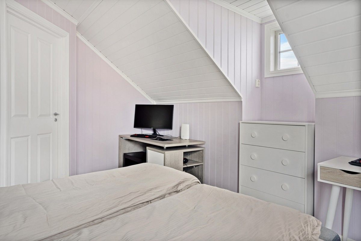 A bedroom with a sloped ceiling, a bed, a dresser, a desk with a computer, and a window.