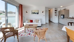A modern living room with a sofa, a coffee table, wicker chairs, and a kitchen area with cabinets and appliances.