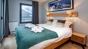 A neatly made bed with a green blanket, two bedside lamps, a framed picture on the wall, and a window with curtains. - La Cordee 124 Apartment -  an apartment sleeping 6 with parking & pool (Chamonix-Mont-Blanc)