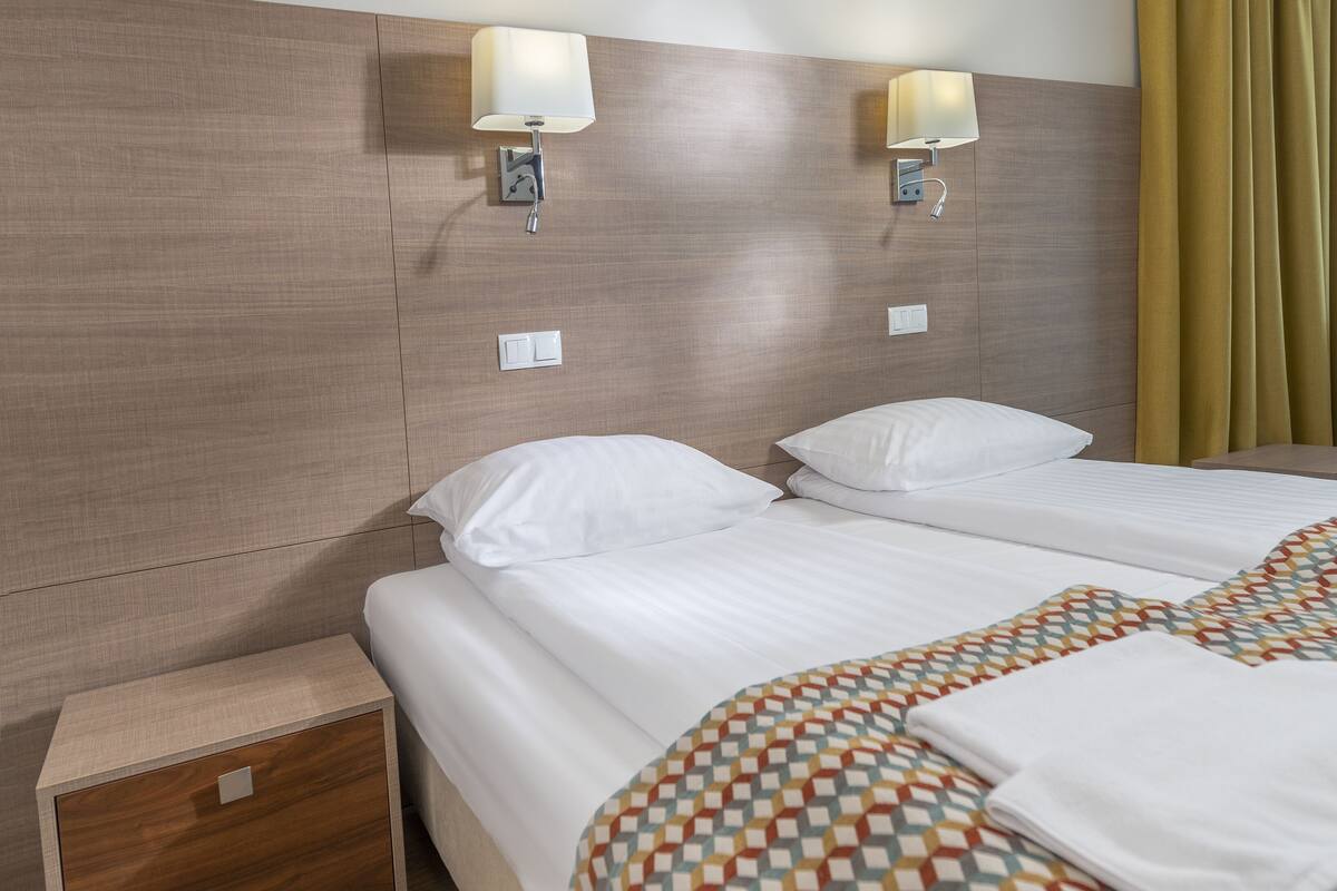 A hotel room with two beds, a bedside table, and a wall-mounted lamp.
