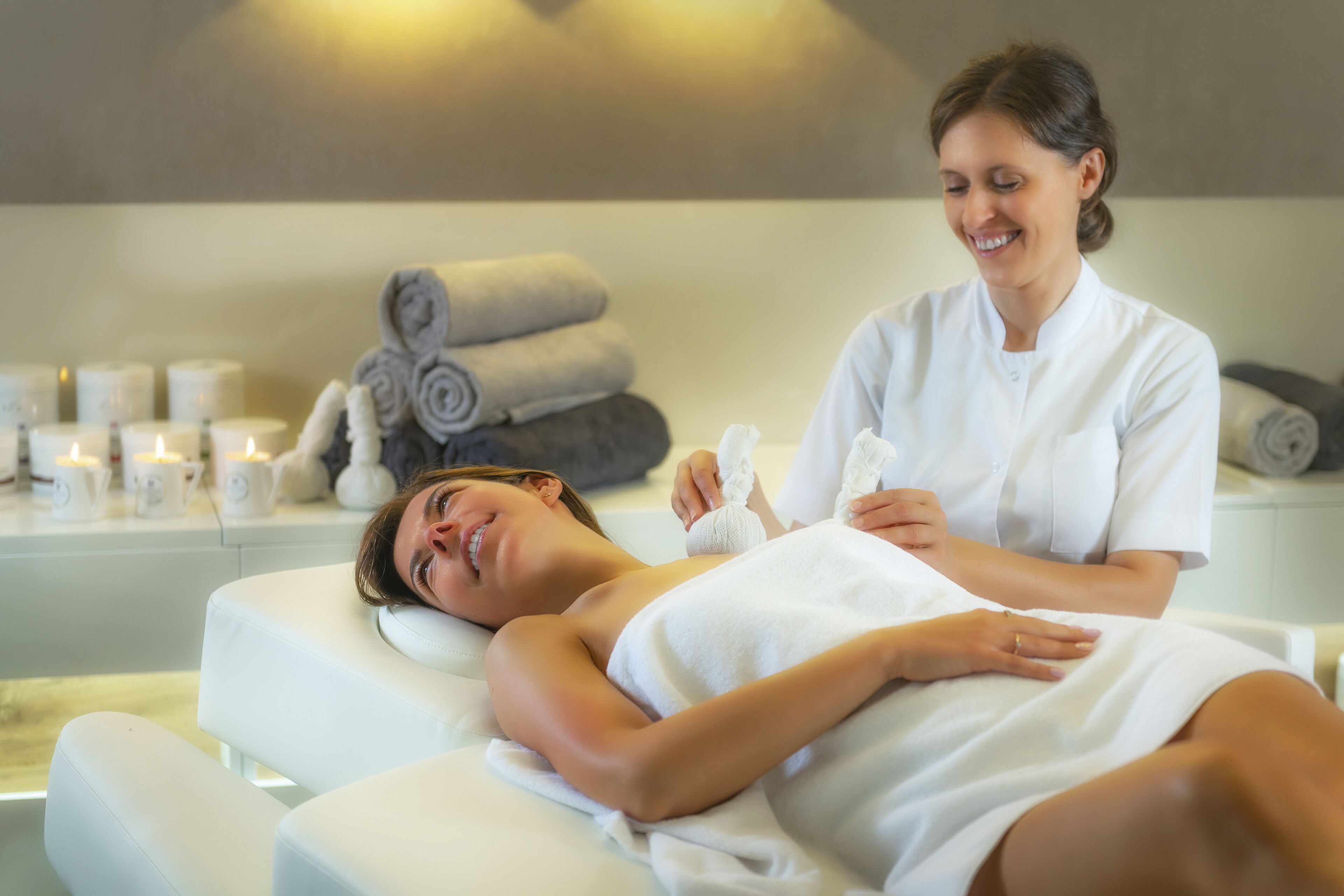 A person lying on a massage table in a spa setting with towels and candles in the background.