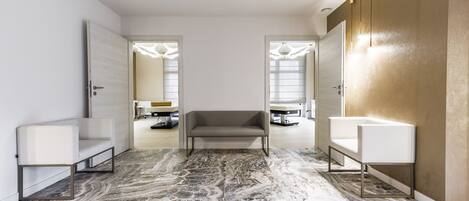 Una habitación de hotel moderna con suelo de mármol, un sofá gris, un sillón blanco y un espejo grande.
