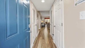 Un pasillo con una puerta azul identificada con el número '202', puertas blancas, suelo de madera y un comedor al fondo.