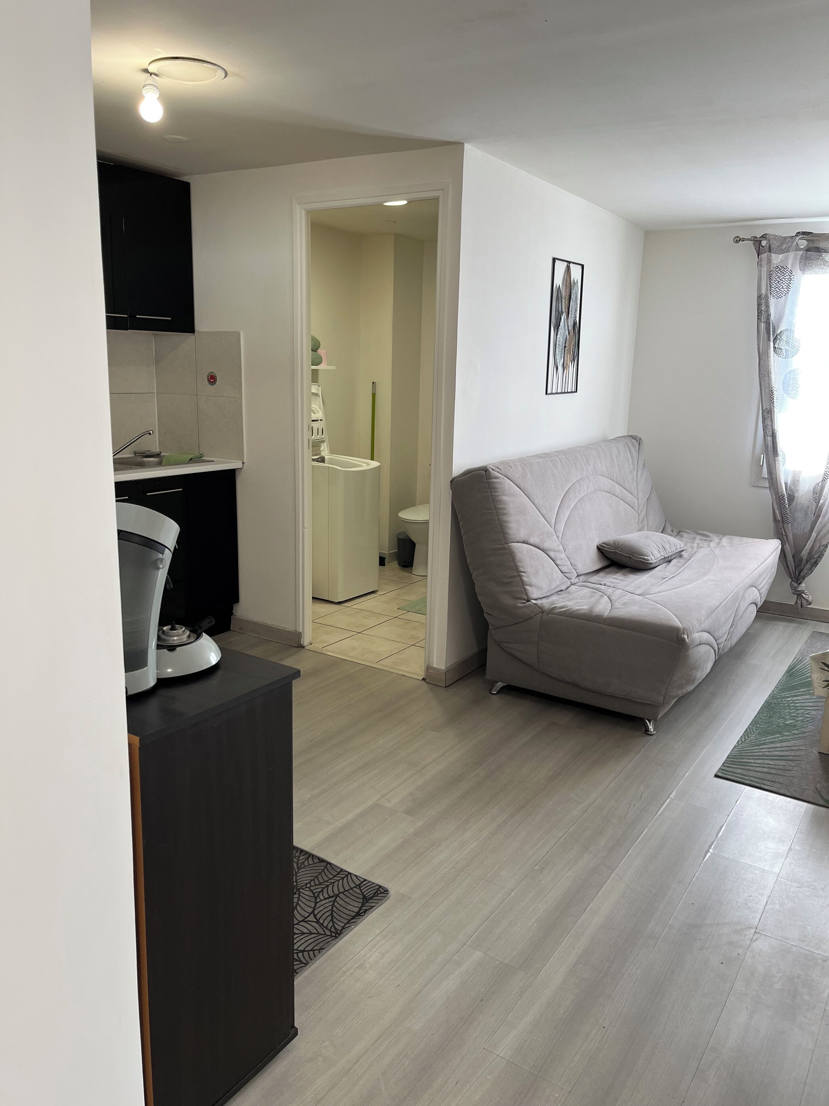 Een moderne woonkamer met een grijze bank, een klein keukenhoekje en een badkamer zichtbaar door een open deur.