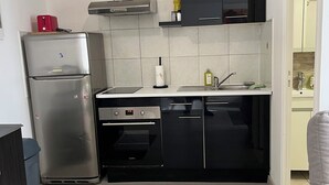 Ett kompakt kök med ett kylskåp i rostfritt stål, en inbyggd ugn och en mikrovågsugn. Här finns en diskho med blandare och en bänkskiva med en flaska diskmedel.