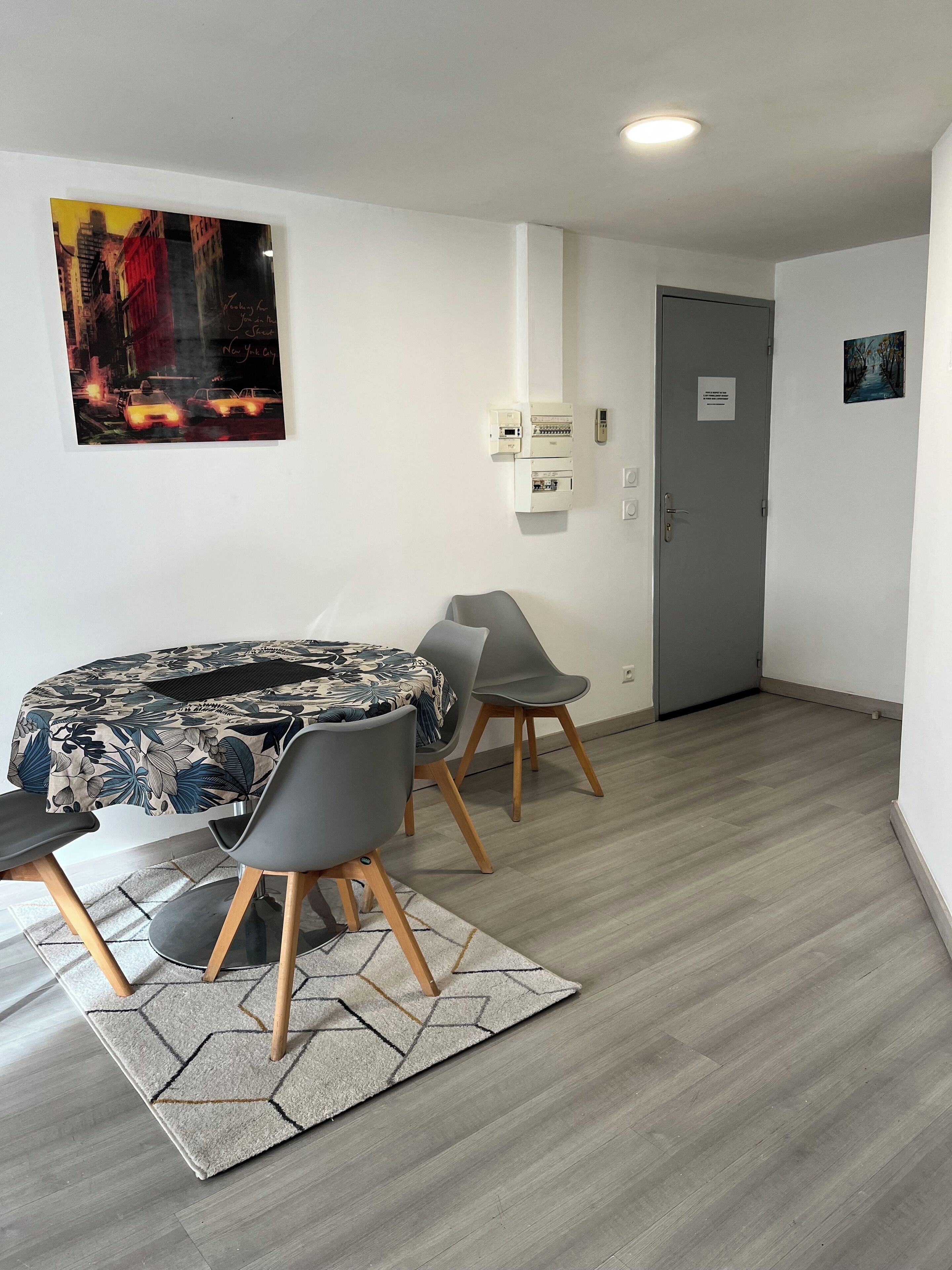 Une pièce moderne avec une table ronde, deux chaises grises, une nappe à motifs floraux, une porte grise et un tableau représentant un paysage urbain.