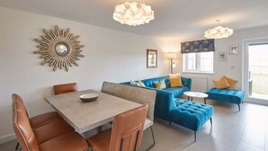 A modern living room with a blue sofa, a dining table with chairs, and a decorative sunburst wall piece.