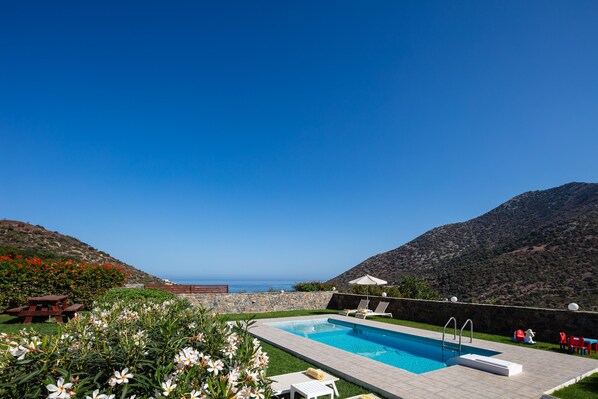 Relax by the serene pool surrounded by lush greenery and vibrant flowers, with a scenic sea and hill view.