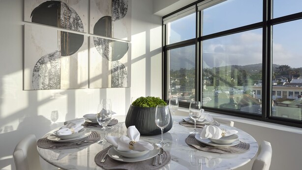 Comedor con una mesa redonda para cuatro, una planta en maceta y vistas de la ciudad a través de amplias ventanas.