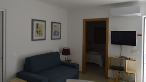 Ett vardagsrum med en blå soffa, en tv som är monterad på väggen och en dörr som leder till ett sovrum.