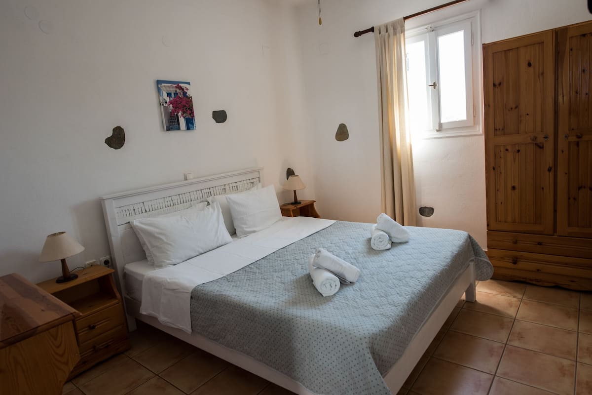A neatly made bed with white linens and two rolled towels, a wooden nightstand with a lamp, a small picture on the wall, and a wooden wardrobe.