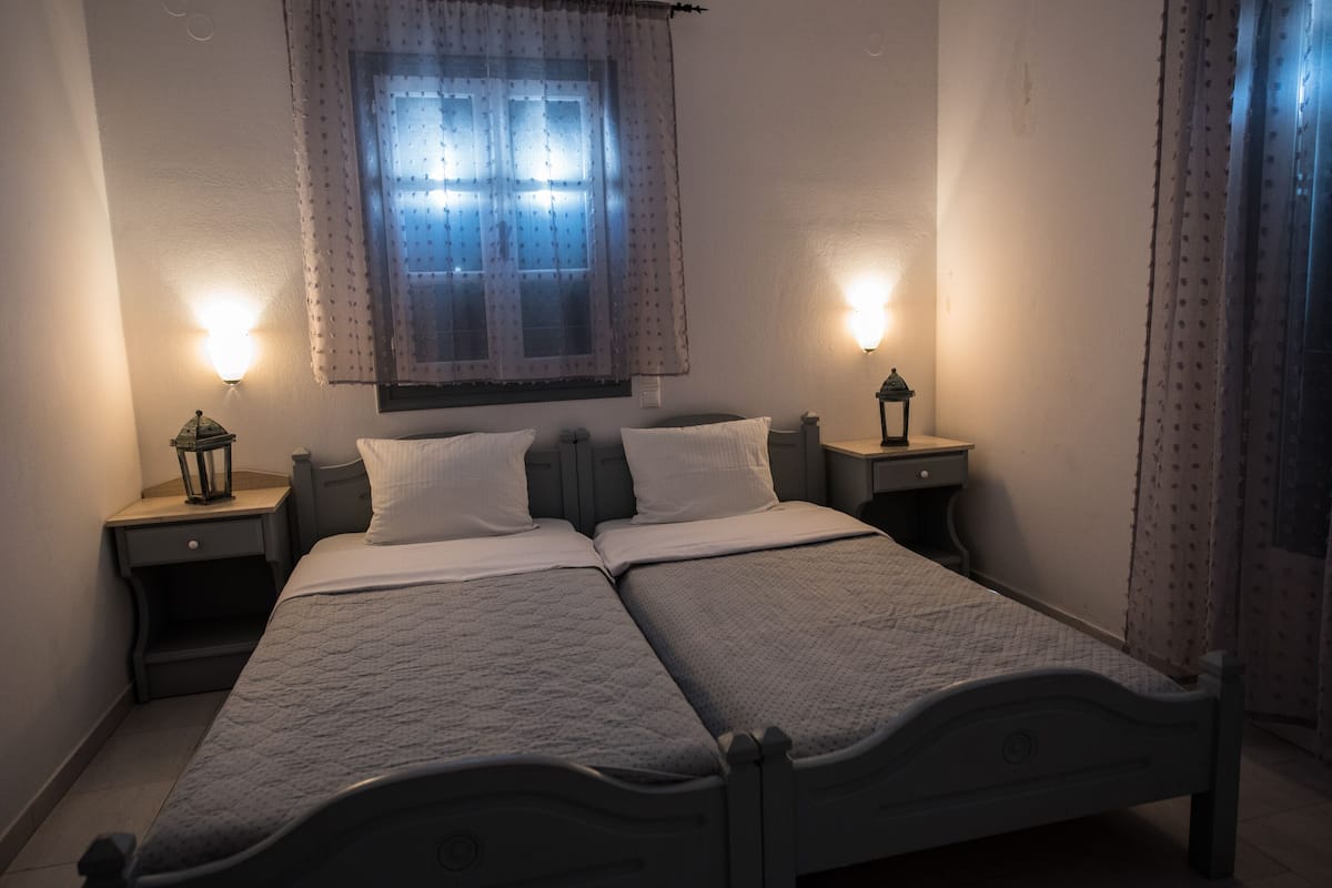 A double bed with a quilted cover, two bedside tables with lamps, a window with a patterned curtain, and a wall-mounted light fixture.