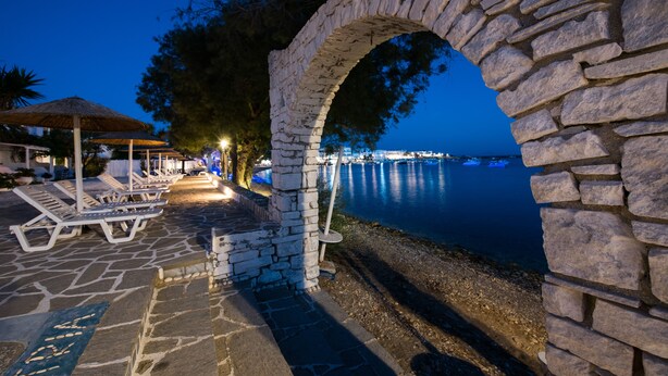 A stone archway leading to a beachside area with lounge chairs and umbrellas, overlooking calm waters. - Ostria Studios & Apartments (Paros)