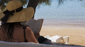 A person reading a book on a beach with a straw hat, a beach chair, and a tree in the background. - Ostria Studios & Apartments (Paros)