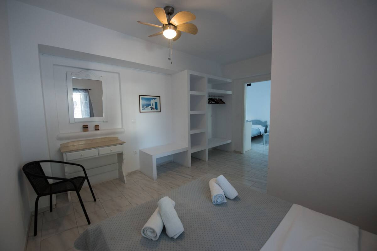 A modern, minimalist bedroom with a bed, a desk, a chair, a ceiling fan, and a view of the outside.