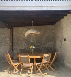 An outdoor dining area with a round wooden table and six chairs, a woven light fixture, and a stone wall backdrop. - Peaceful house in the heart of the Pyrenees (Igon)