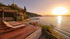 Eine Holzterrasse mit Liegestühlen und einer Sitzecke mit Blick auf ein Gewässer bei Sonnenuntergang.