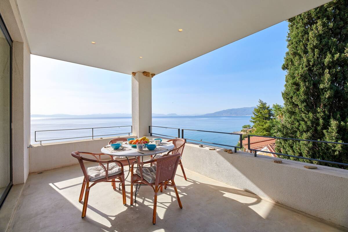 A balcony with a table set for dining, overlooking a scenic view of the sea and mountains.