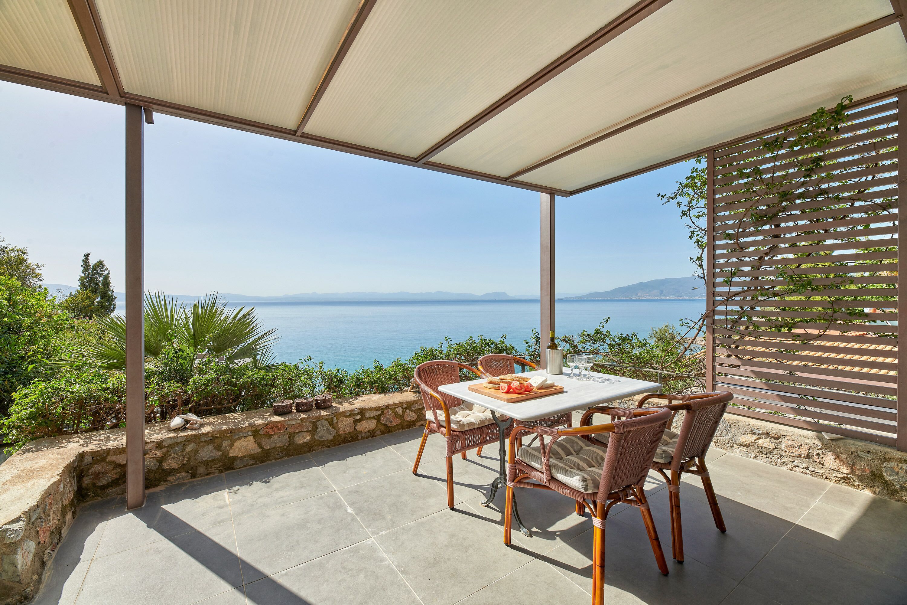 A covered outdoor seating area with a table and chairs, overlooking a scenic view of the sea and hills.