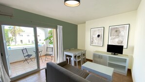 Un salon moderne avec un canapé, une table à manger et un téléviseur. Il y a un balcon avec des chaises et une vue sur un immeuble et des arbres.