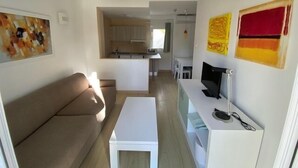 Ett modernt vardagsrum med en soffa, ett soffbord och en tv som står på ett tv-bord. I bakgrunden finns ett kök med skåp och ett fönster.