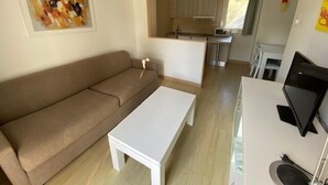 Ett vardagsrum med en brun soffa, ett vitt soffbord och en tv som står på ett tv-bord. Det finns också en köksdel med skåp och en mikrovågsugn.